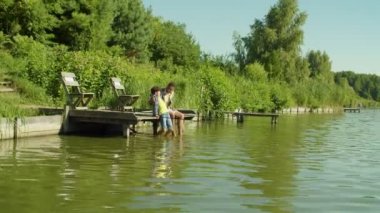 Positive African father and happy adorable multiethnic teenage son sitting in embrace on wooden jetty on lake, dangling feet into water, bonding and enjoying scenic nature during fishing.