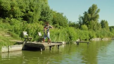 Loving black father teaching lovely school age multicultural son to cast spin fishing rod from wooden pier on lake, showing support and care while multiethnic family enjoying angling on summer day.