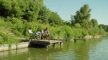 Excited joyful adorable multiracial teenage fisherman catching fish with spin fishing rod and happy African father helping with landing net, while family enjoying angling from wooden pier on lake.