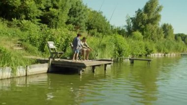 Attractive African father twisting spin fishing rod and excited cute school age multiethnic son helping with landing net to catch fish, while joyful family enjoying fishing from wooden jetty on lake.