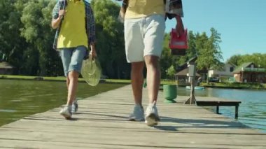 Low section of multiracial family with preadolescent son holding fishing rods, tackle box and equipment stepping on wooden pier, going for fishing on pond on summer day.