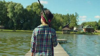 Rear view of positive teenage fisherman with fishing tackle box carrying fishing rod on shoulder walking for fishing along wooden pier on lake, enjoying recreational activity on summer day.