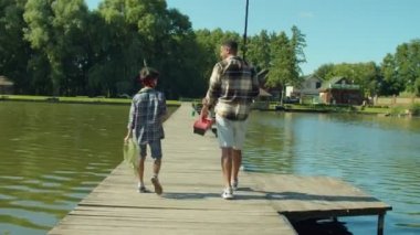 Rear view of happy school age multiracial boy and caring black father with fishing tackle box and equipment carrying fishing rods, communicating and enjoying leisure while walking for fishing on lake.