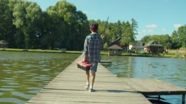 Back view of cute teenage fisherman with fishing tackle box carrying fishing rod on shoulder, walking for fishing on pond along wooden pier while enjoying leisure on summer day.