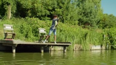 Skillful adorable Arab school age fisherman fishing from wooden jetty on pond, casting spin fishing rod and enjoying leisure and scenic summer nature during weekend.