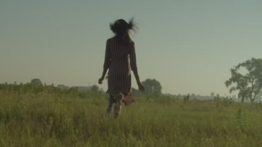 Rear view of joyful attractive African American woman in summer dress running through green fields, enjoying recreational activity and scenic nature, expressing carefree mood and happiness at dawn.