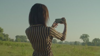 Rear view of cheerful attractive African woman tourist standing on green grass field, photographing scenic summer nature on cellphone while enjoying recreation in countryside at sunrise.