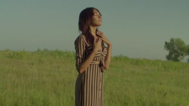 Portrait of serene charming black female in sundress with eye closed standing on green grass field, inhaling fresh air, enjoying recreation and scenic nature while resting in countryside .