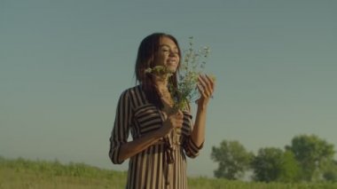Relaxed charming African American female in sundress with eyes closed standing on meadow, enjoying aroma of fresh blossom wildflowers, expressing happiness while relaxing in summer nature.