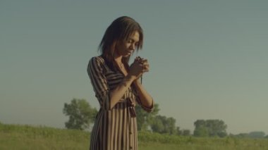 Portrait of Christian beautiful African American woman with eye closed holding catholic rosary, saying prayers, asking for bless, mercy and peace while standing on green field in morning