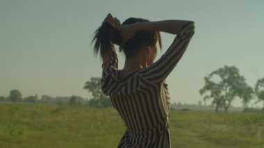 Portrait of serene charming black female in sundress enjoying leisure and scenic summer nature, relaxing in rays of warm sunrise while standing on grass field during vacations in countryside.