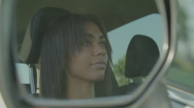 Reflection of charming joyful African female driver looking with radiant smile in car side rear view mirror, expressing positivity and happiness while driving automobile.