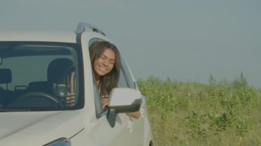 Carefree cute African American female driver leaning out car window, waving hello gesture, showing happiness, friendliness and cheerful mood while enjoying road trip on summer vacations in countryside