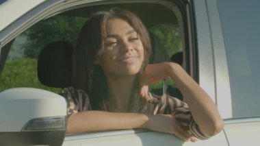 Happy attractive African American woman traveler sitting in drivers seat, leaning on car window, looking with cheerful smile while enjoying scenic summer nature during road trip in countryside.