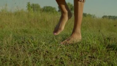 Close-up of beautiful barefoot black female legs gracefully stepping on green grass in rays of rising sun during summer vacations in countryside.