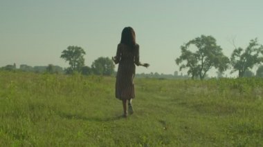 Rear view of elegant attractive African female traveler in sundress walking through green grass field, enjoying summer vacations and beauty of nature while spending leisure in morning in countryside.