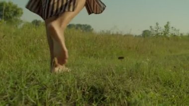 Close-up of slim barefoot black female legs gracefully walking on green grass field in ray of warm summer sunrise. Side view.
