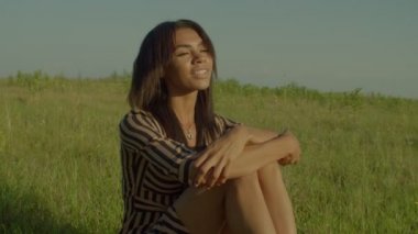 Happy elegant attractive black woman tourist sitting on green grass field, enjoying leisure and scenic summer nature in morning , expressing cheerful mood and positivity while resting in countryside