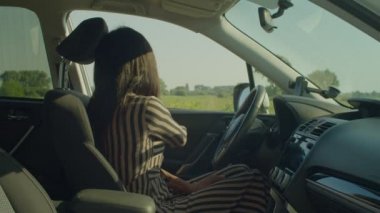 Side view of confident charming African American female driver in sundress buckling up car seatbelt, holding steering wheel in rays of warm rising sun over scenic nature during road trip.