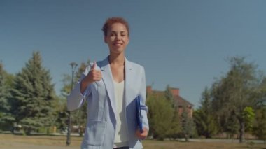 Başarılı, kıvırcık saçlı Afrikalı Amerikalı iş kadını elinde klasör, elinde jest, dışarıda dururken pozitifliğini ve mutluluğunu ifade ediyor..