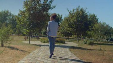 Yüksek topuklu ayakkabılar ve resmi kıyafetler giyen, klasör ve tablet bilgisayarı taşıyan, yaz günü şehir parkında yürürken iş görüşmelerine giden şık, çekici Afrikalı Amerikalı iş kadınının arka görünüşü..