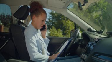 Resmi giyimli, başarılı Afrikalı Amerikalı iş kadını sürücü koltuğunda oturuyor, iş arkadaşlarıyla cep telefonuyla konuşuyor ve yolculuk sırasında belgelerle uğraşıyor..