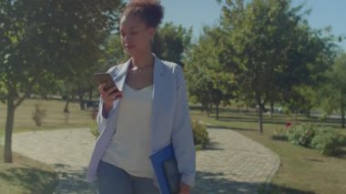 Parkta yürürken iş ortaklarıyla internet üzerinden mesajlaşmak, dijital tablet ve belgeler taşıyan başarılı siyahi kadın yönetici..