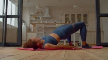 Motivasyonlu, aktif giyimli, seksi kadın vücut ağırlığı kalça köprüsü egzersizi yapıyor, kalça kaslarını güçlendiriyor ve evdeki eğitim süresince vücut kaslarının dengesini arttırıyor..