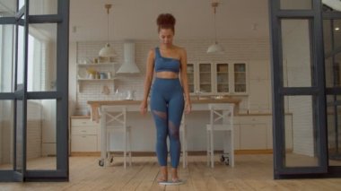 Neşeli, pozitif, aktif giyimli, kilolarını ölçen güzel bir Afrikalı kadın, duygusal olarak kilo verme başarısını jestler kazanarak kutluyor, evde mutluluk ve heyecan ifade ediyor..