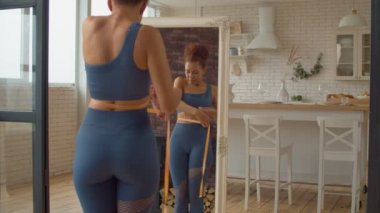 Aktif giyimli, pozitif, neşeli ve çekici siyahi kadının yansıması kilo verme başarısını kontrol ediyor, ölçüm bandıyla belini ölçüyor ve içeride diyet yaptıktan sonra vücut şekline hayran kalıyor..