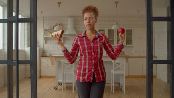 健康とジャンクフードの間で選択美しいアフリカ系アメリカ人女性を痩身 意思決定を行い 国内の部屋に立っている間 おいしい新鮮なリンゴをかむ — ストック動画