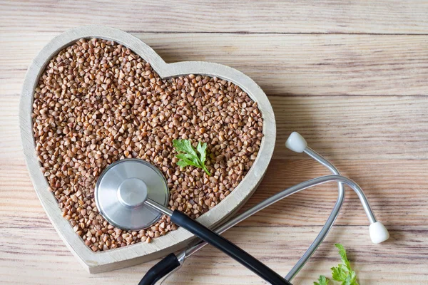 Buckwheat in heart-shaped bowl with stethoscope on wooden background, healthy heart food concept