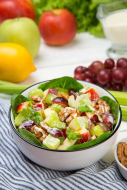 Waldorf salad of apples, celery stalks, walnuts, grapes, lettuce in a white salad bowl with ingredients on an old rustic white wooden table. Selected focus.
