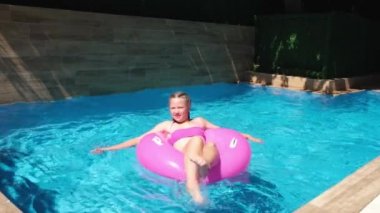 Turkuaz suyla yüzme havuzunda dinlenen bir kız. Pembe şişme yüzükle yüzen kız, ayaklarıyla su sıçratıyor. Seyahat ve tatil kavramı