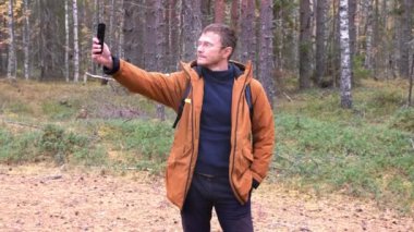 Neşeli turist sonbahar ormanında selfie çeker ve dans eder. Ormanda yürürken akıllı telefonlu bir adam.