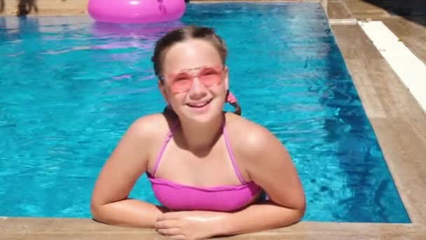 Chica adolescente sonriente en gafas de sol de color rosa en la piscina. Chica sentada en el borde de la piscina. concepto de vacaciones soleadas calientes — Vídeo de stock
