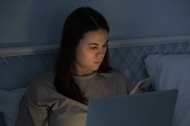 Woman using laptop computer and surfing social media on smartphone in bed before sleep at night. Insomnia, sleep disorder concept