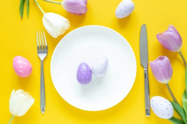 Paskalya sofrası. Beyaz tabak, dekoratif paskalya yumurtaları, laleler ve sarı arka planda gümüş çatal bıçak takımı.