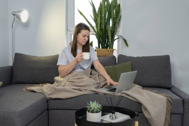 Evdeki kanepede otururken elinde kupa olan genç bir kadın dizüstü bilgisayar kullanıyor.