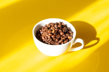 Sarı arka planda beyaz seramik fincanda kahve çekirdekleri. Sert güneş gölgeleri