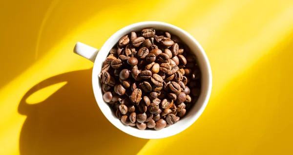 Granos de café en una taza de cerámica blanca sobre un fondo amarillo. Sombras de sol duras. Vista superior — Foto de Stock