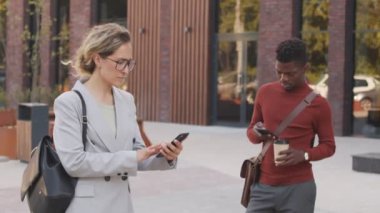 Şehir merkezindeki açık havada akıllı telefonlardan kayan beyaz kadın ve Afro-Amerikan erkek iş kadınlarının orta yavaş çekimi.