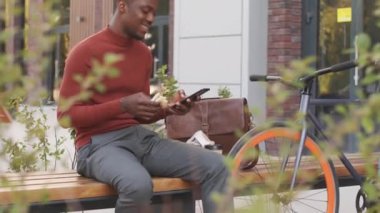 Akıllı günlük giyimli, neşeli, Afro-Amerikan bir adamın akıllı telefondan kayarak öğle yemeği molası verirken ve modern iş bölgesinde bankta oturup sandviç yerken yavaş çekim.