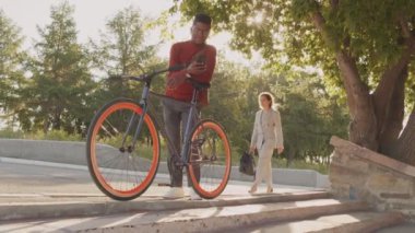 Akıllı günlük giyimli ve gözlüklü genç bir Afro-Amerikalının güneş ışığının altında bisiklet sürerken, akıllı telefonda kayarken ve gülümserken çekimleri.