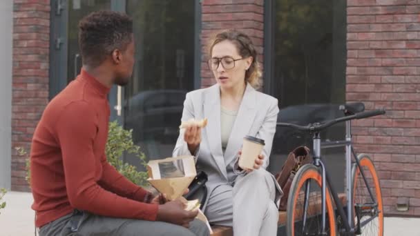 市内中心部のベンチに座ってサンドイッチを食べながら 屋外で一緒に昼食を取っている現代的な多民族ビジネスカップルの中低速ショット — ストック動画