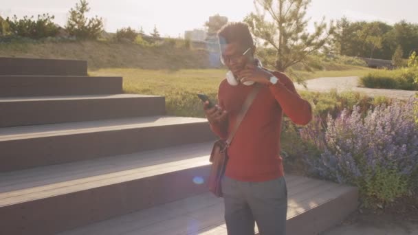 在阳光明媚的日子里 身穿红色高领衫的非洲裔美国年轻人打开智能手机 戴上耳机站在城市公园里 拍了一张中等速度的照片 — 图库视频影像