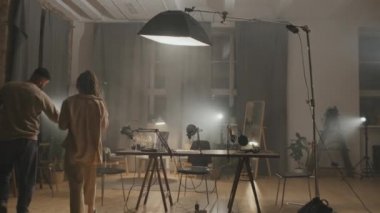 Genç bir kadın ve erkeğin evde oturmuş podcastlarını kaydetmeye hazırlandıkları orta boy bir fotoğraf. Odanın etrafına parlak stüdyo ışıkları yerleştirildi