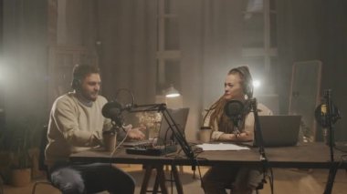 Akşamları oturma odasındaki masada oturan ve online radyo programlarını yayınlayan kulaklıklı genç adam ve kadının orta boy fotoğrafları. Mikrofonlara konuşuyor ve gülüyorlar.
