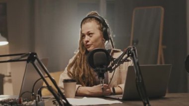 Karanlık oturma odasındaki masada oturan ve mikrofona konuşan neşeli, genç bir bayanı takip etmek ve kamera dışında biriyle konuşmak ve evde podcast yapmak.