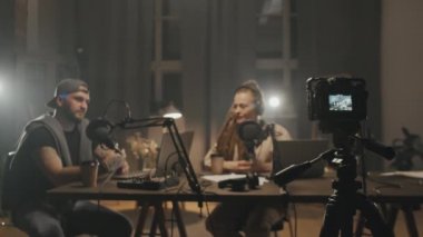 Oturma odasında lamba ve stüdyo ışıklarıyla aydınlatılan ve podcast 'i kaydeden neşeli adam ve kadının video kamera görüntülerini izliyoruz.